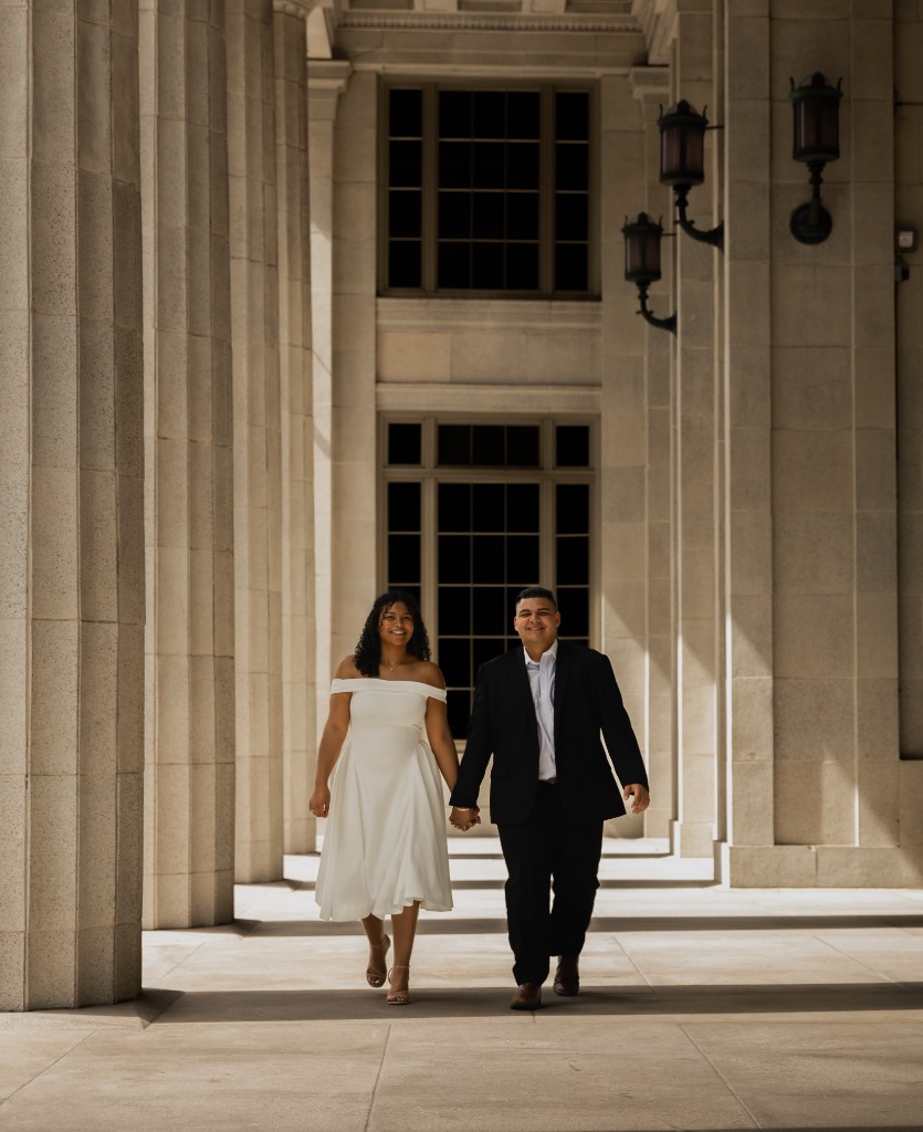 Engagement couple in a black suit and white off-the-shoulder dress holding hands and walking smiling through classical concrete pillars