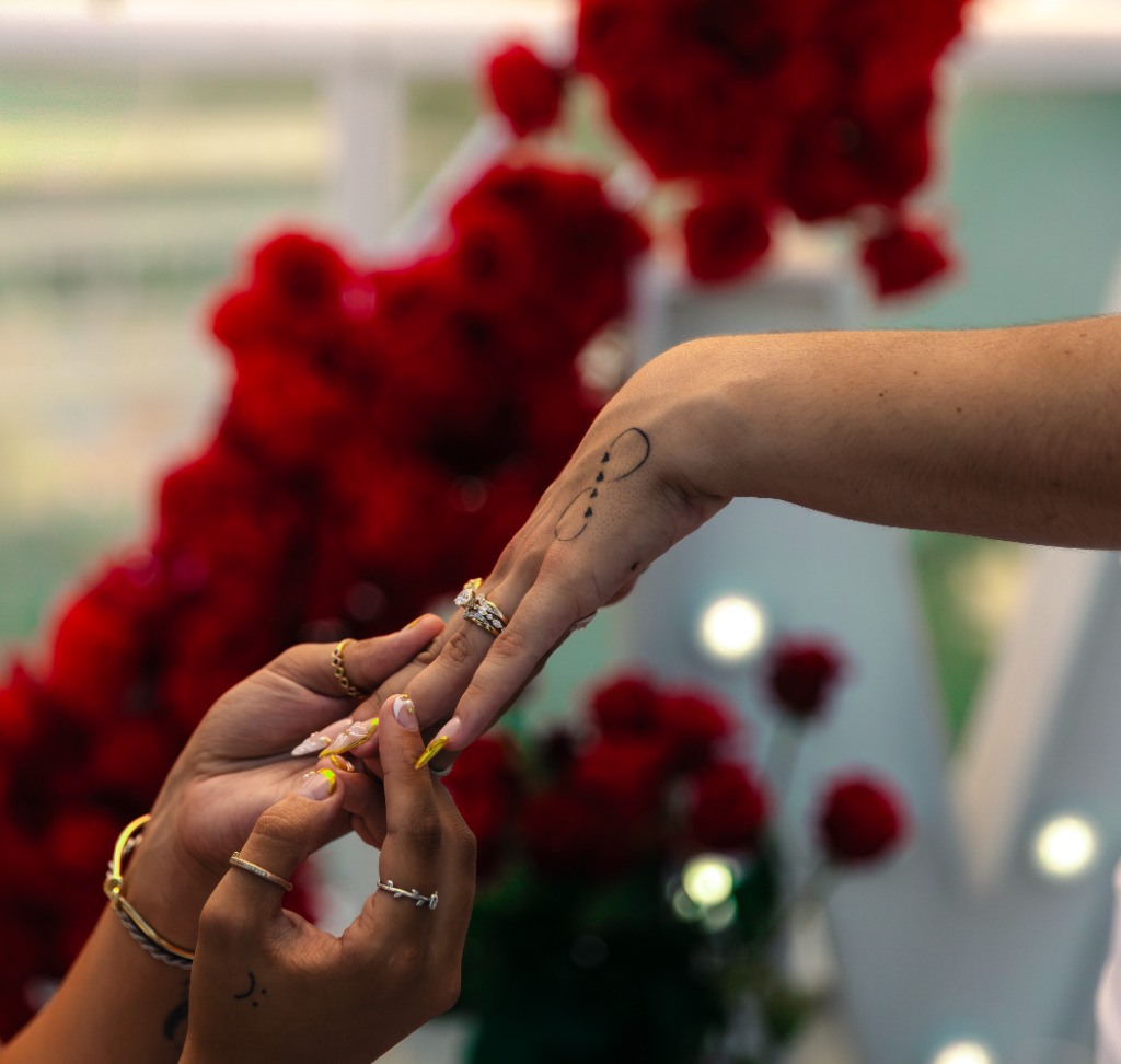 Close-up of one partner's hands gently placing a diamond engagement ring on the other's finger against a soft red rose backdrop