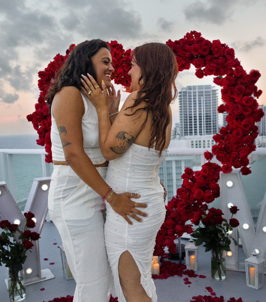 One partner embracing the other from behind, both smiling happily while showing off the diamond engagement ring in front of a red rose heart arch