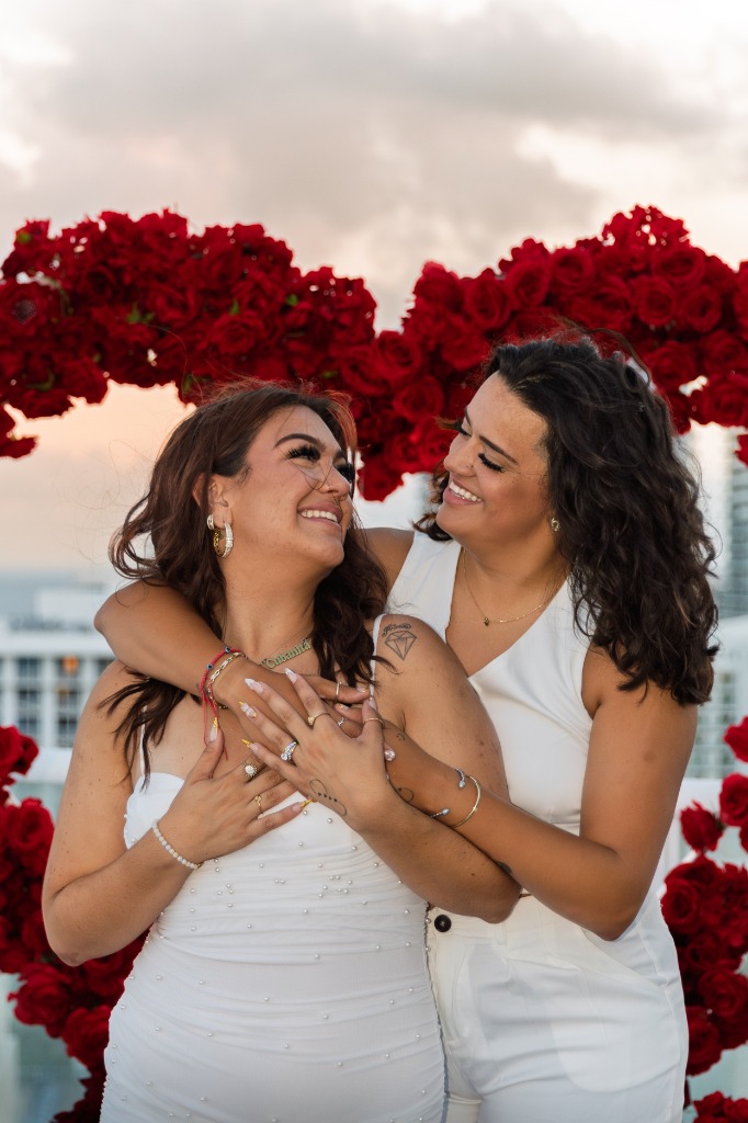 Newly engaged couple embracing face-to-face and smiling warmly in front of a red rose heart archway on a balcony