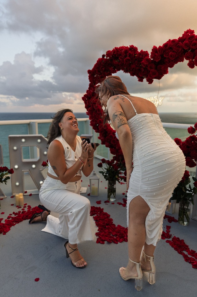 Smiling partner kneeling to propose with a ring box to their laughing, joyful fiancé in front of a red rose heart arch on a rooftop balcony