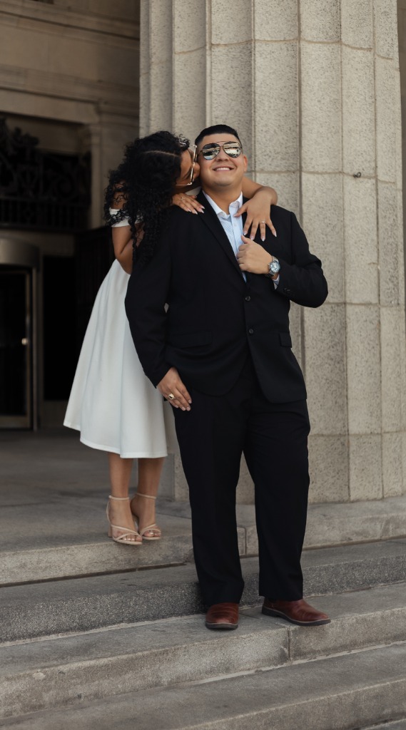 Woman kissing her fiancé's cheek from behind as they stand on classical building steps in front of stone columns