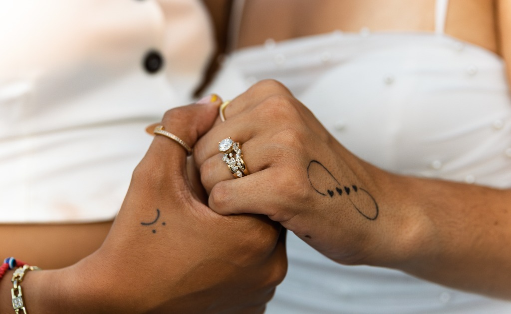 Close-up of the couple's intertwined hands, showcasing a beautiful diamond engagement ring and matching infinity and smile/frown tattoos