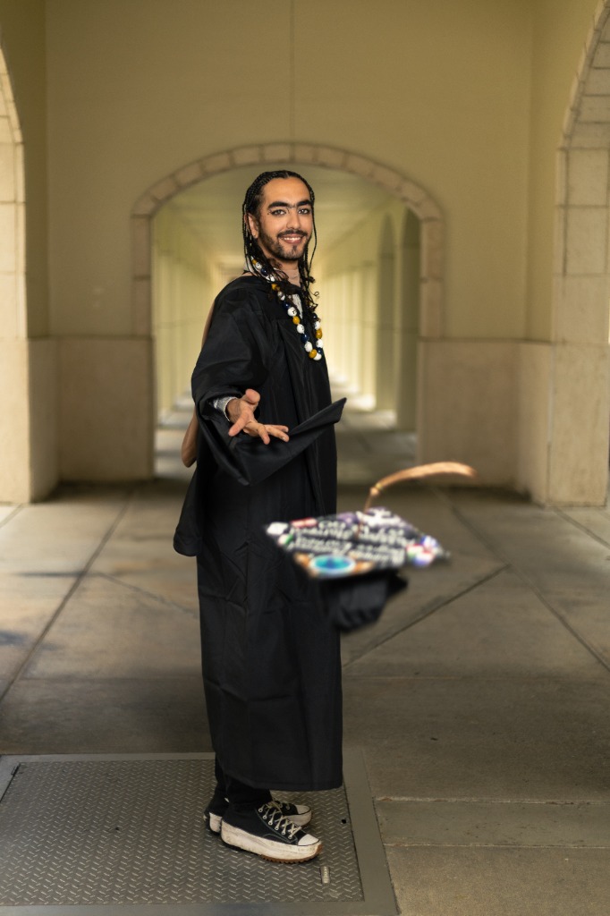 Smiling graduate in black gown tossing graduation cap in air down a beautiful arched corridor
