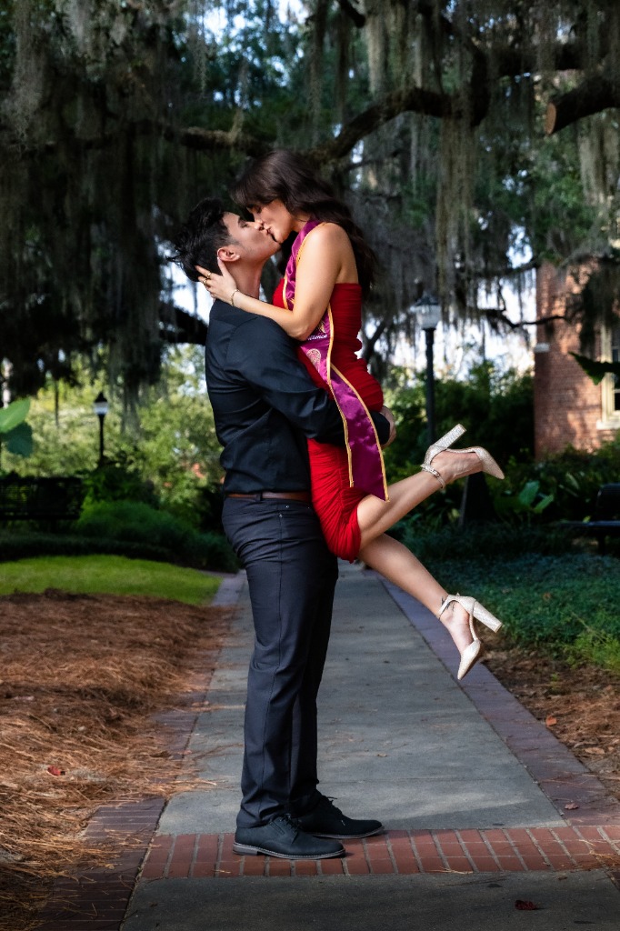 Graduate in red dress and sash being lifted by her partner for a kiss on a garden brick pathway under hanging Spanish moss