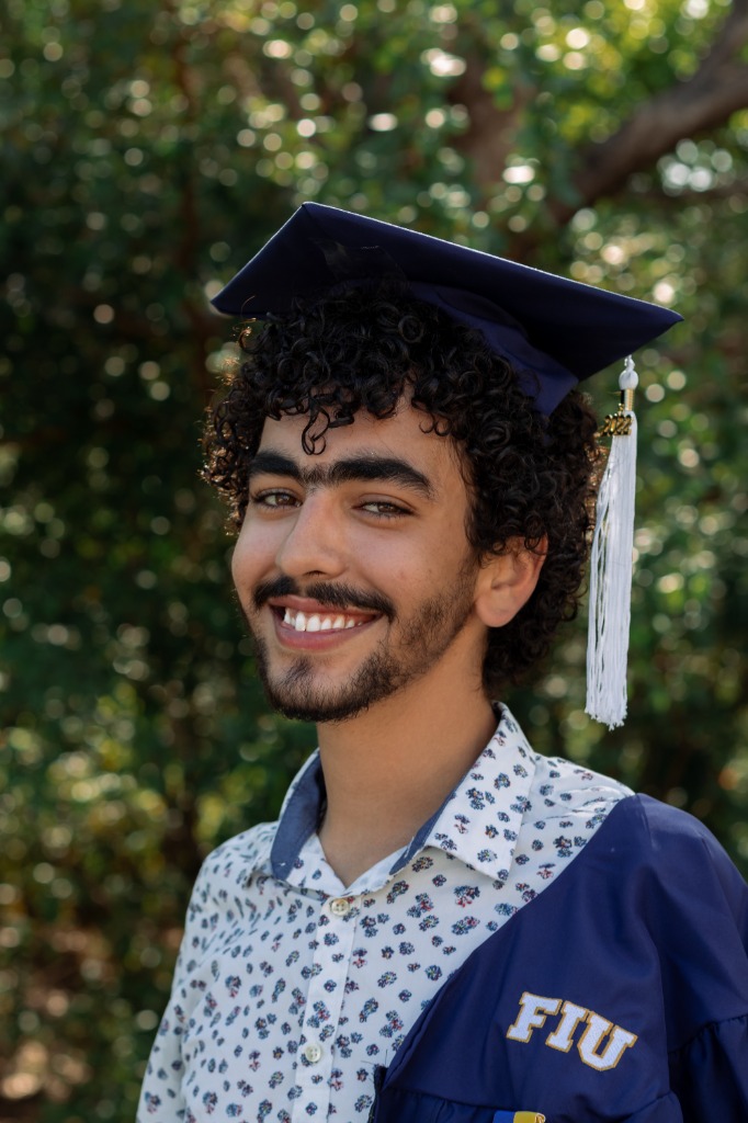 Close-up portrait of a smiling male graduate in cap and gown with a blue FIU sash