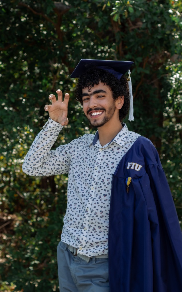 Smiling graduate in cap and gown showing a panther claw hand gesture in front of green trees