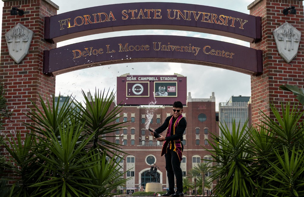 Graduate in FSU sash popping a bottle of champagne under the Florida State University DeVoe L. Moore University Center archway