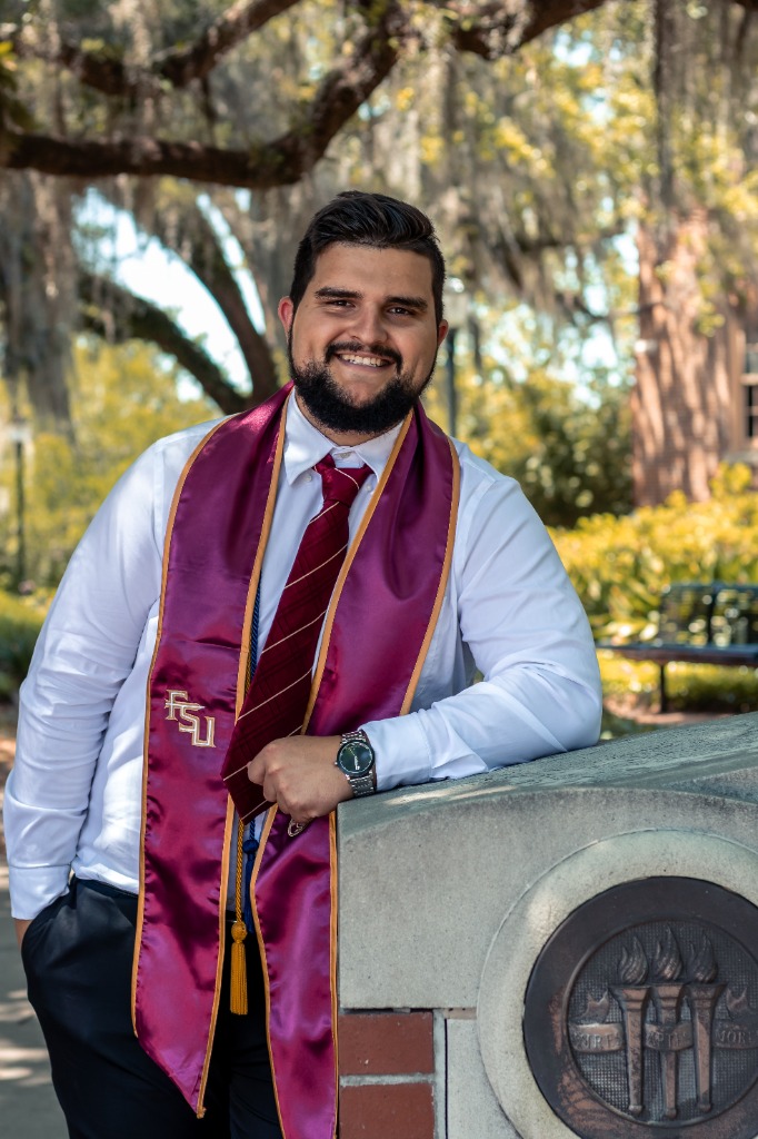 Smiling male graduate in white shirt and FSU sash leaning against a concrete block featuring the FSU seal