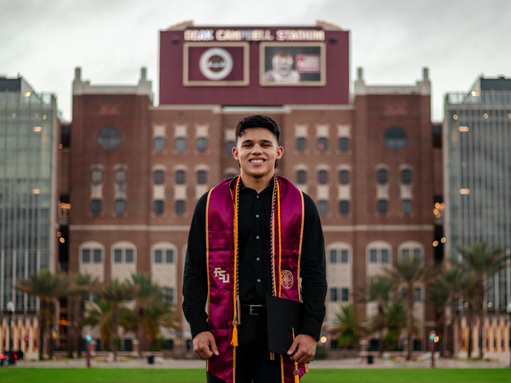 Smiling graduate in black shirt and FSU sash holding cap in front of the Doak Campbell Stadium