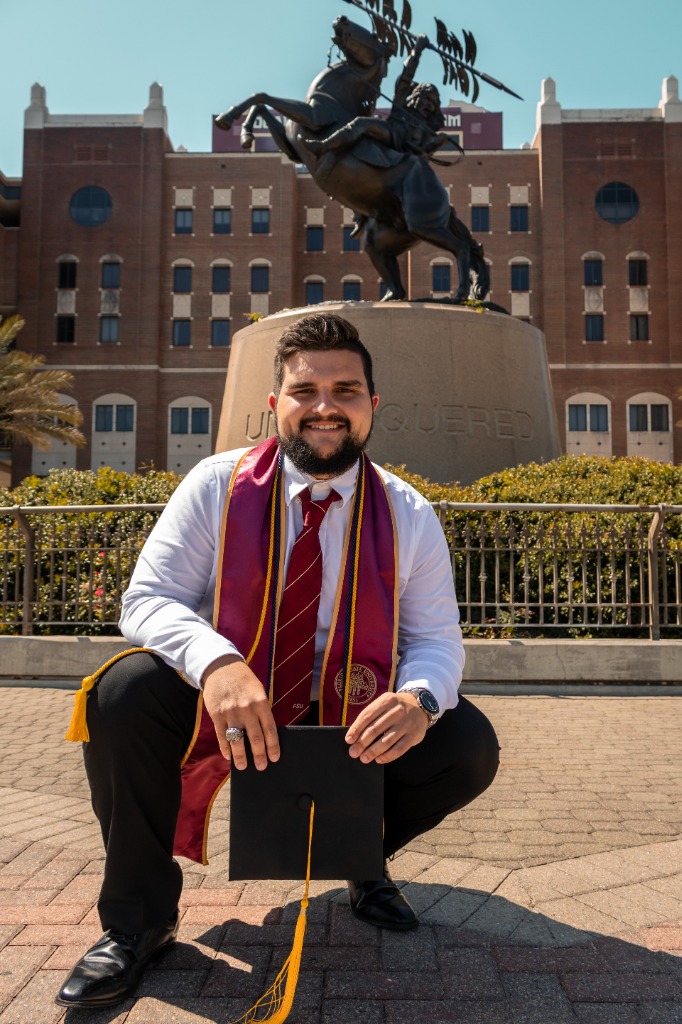 Male graduate in white shirt and FSU sash crouching with his cap in hand in front of the Unconquered statue