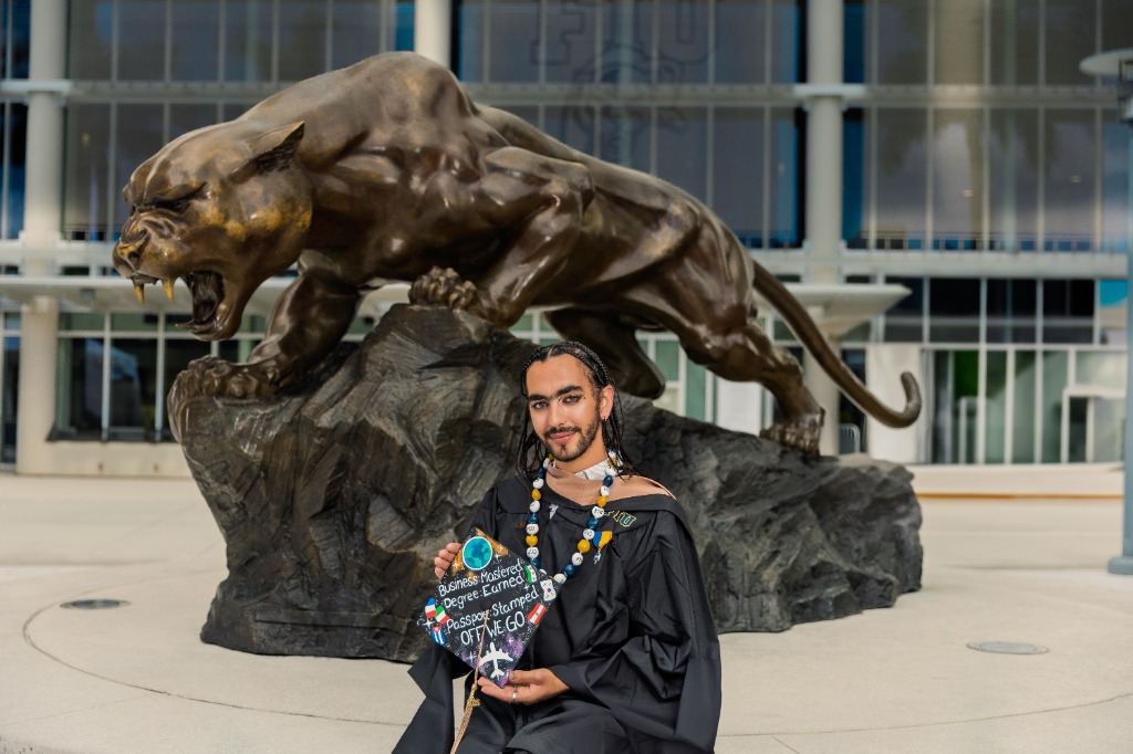 Close-up portrait of a smiling graduate in academic regalia holding their decorated graduation cap next to a bronze panther statue