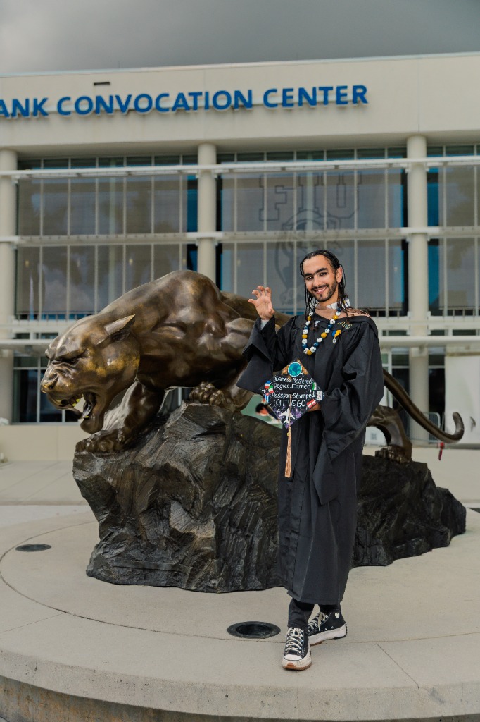 Full-length portrait of a smiling graduate in cap and gown posing proudly next to a large bronze panther statue