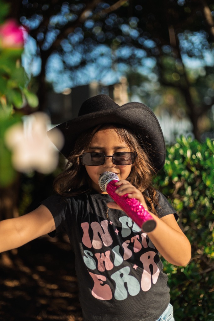 Young girl wearing a cowboy hat and sunglasses singing into a pink glittery microphone