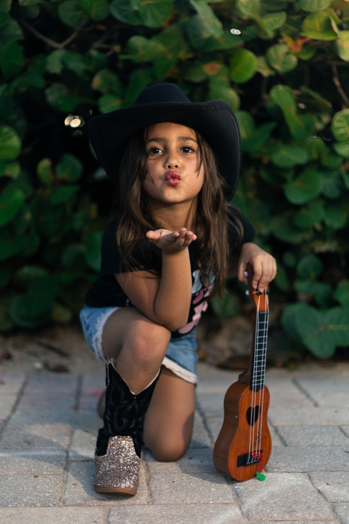 Cute young girl in cowboy hat kneeling next to a ukulele and blowing a kiss