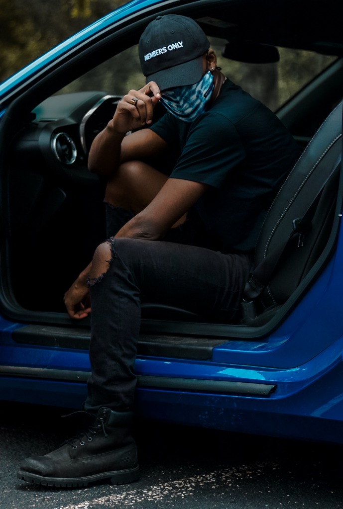 Man wearing a cap and mask sitting inside the open door of a blue car