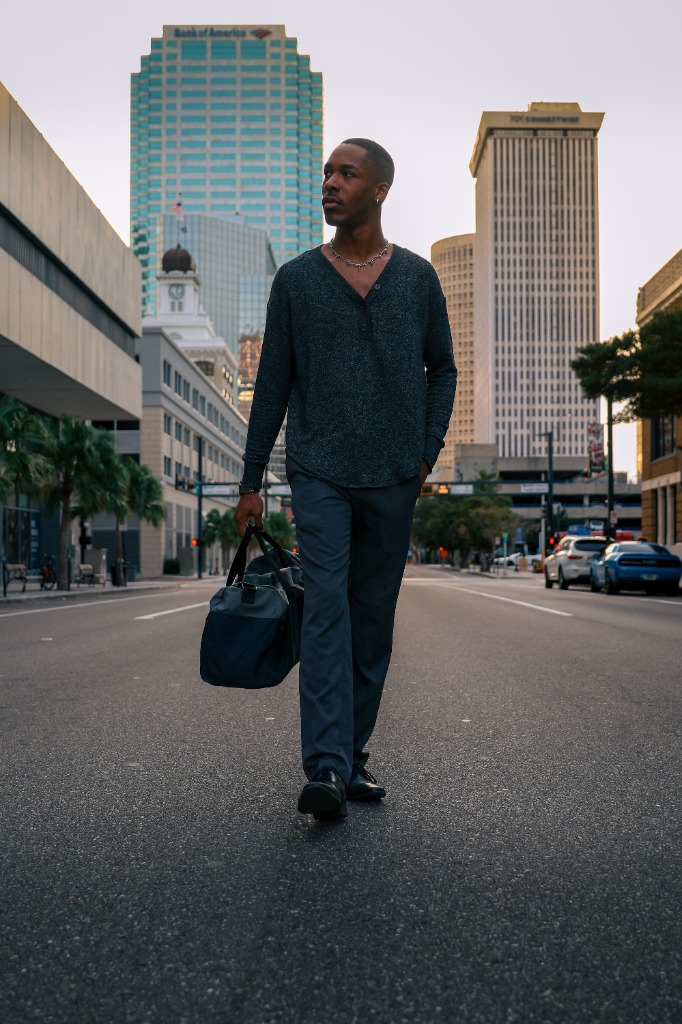 Editorial portrait of a man holding a travel bag walking down a city street with tall buildings behind him