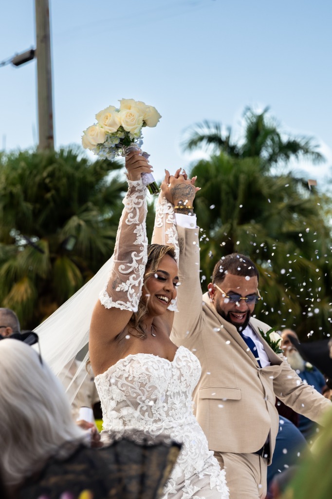 Smiling bride and groom celebrating with arms raised and bouquet high as white confetti rains down